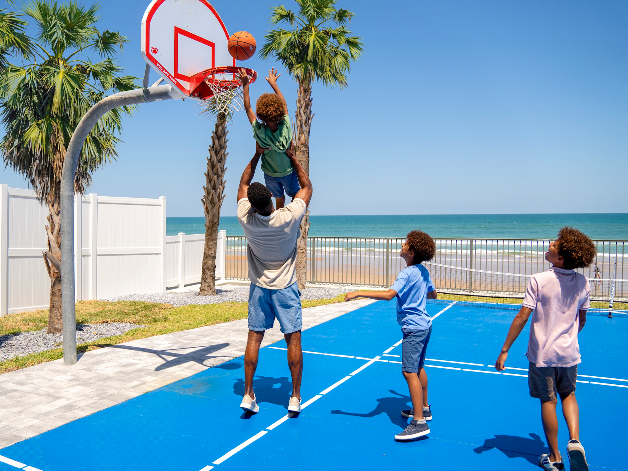 Children playing basketball on an outdoor court with palm trees and ocean view.