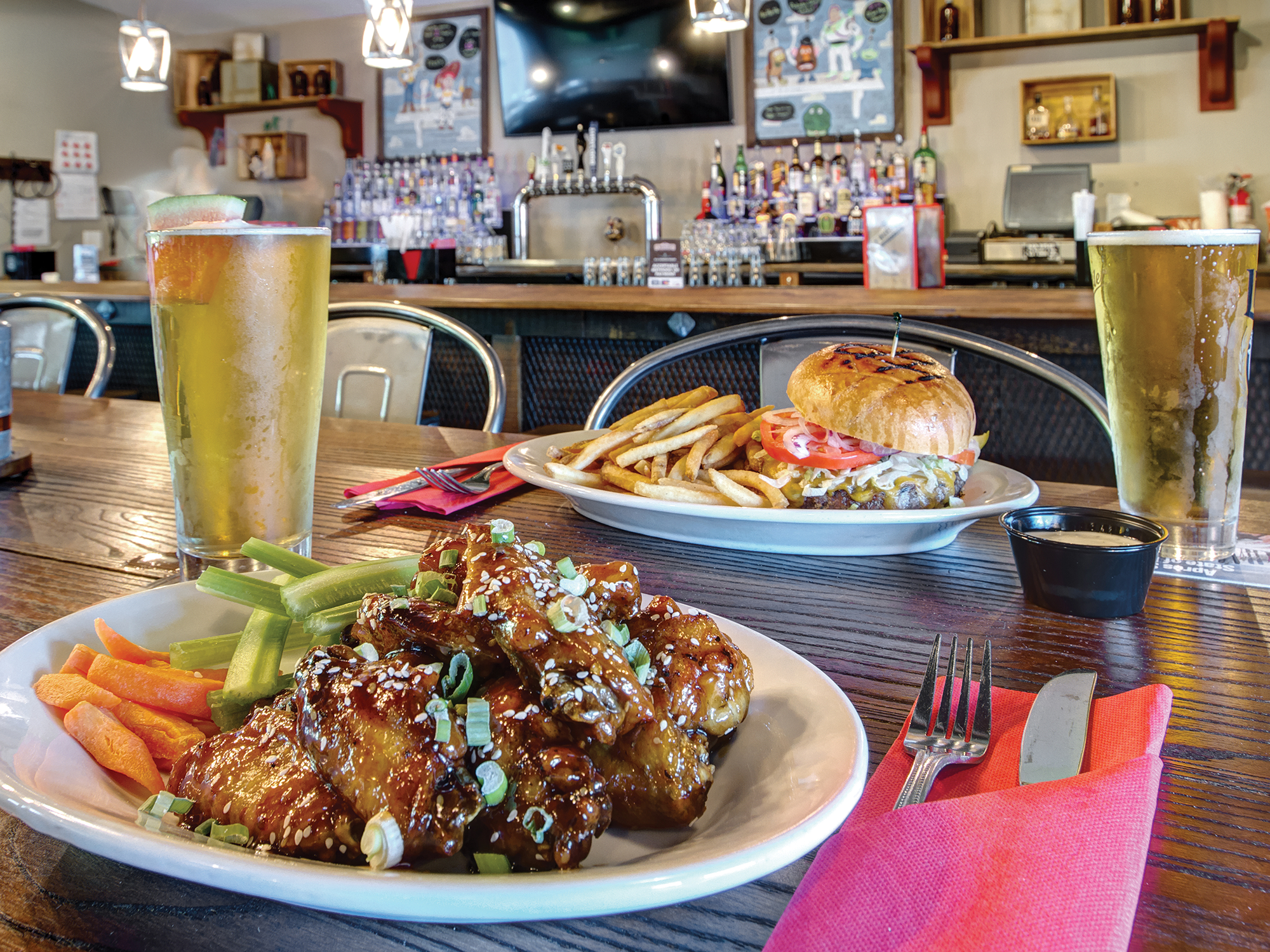 Dinner table with plates of food, including chicken wings and a burger, with drinks and side dishes in a casual dining setting.