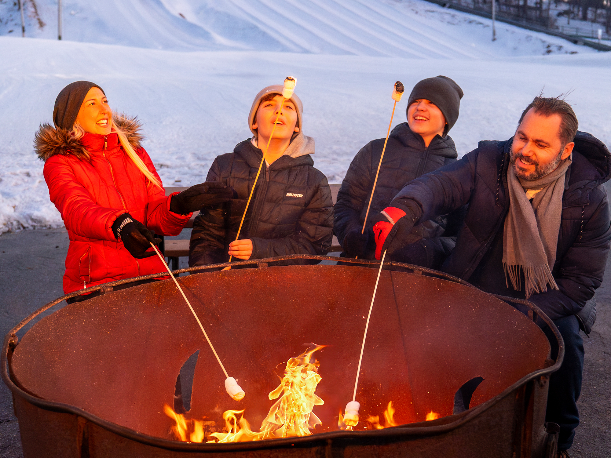 A family of four roasting marshmallows over a fire pit in a snowy landscape.