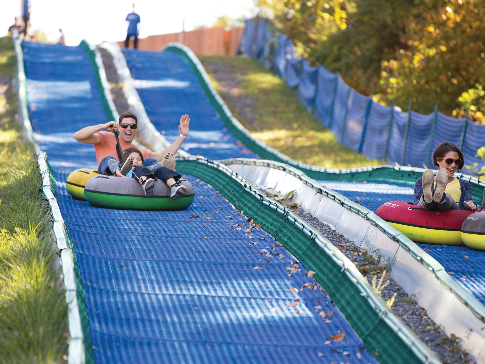 People sliding down a blue slide at an amusement park with green grass and trees in the background.