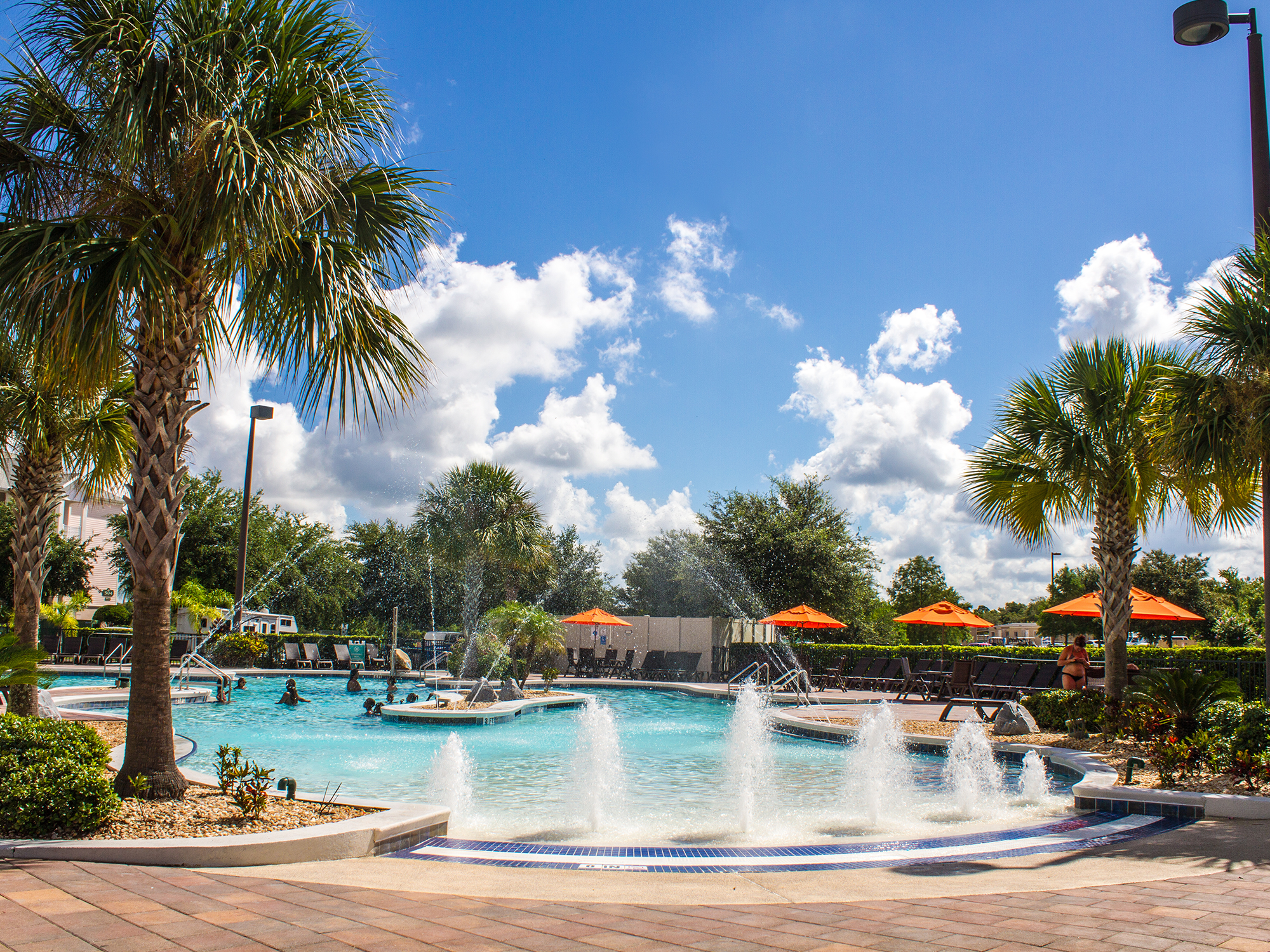 Pool area with fountains, palm trees, and lounge chairs under a blue sky.