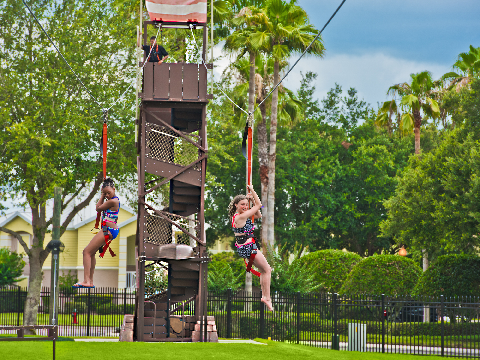 Two kids on a zipline in an outdoor park setting with trees and a clear sky.