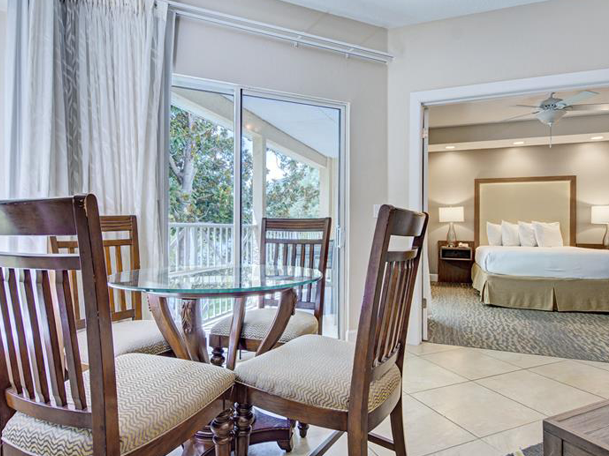 Dining area with wooden chairs and a glass table in a room with a bed and balcony.