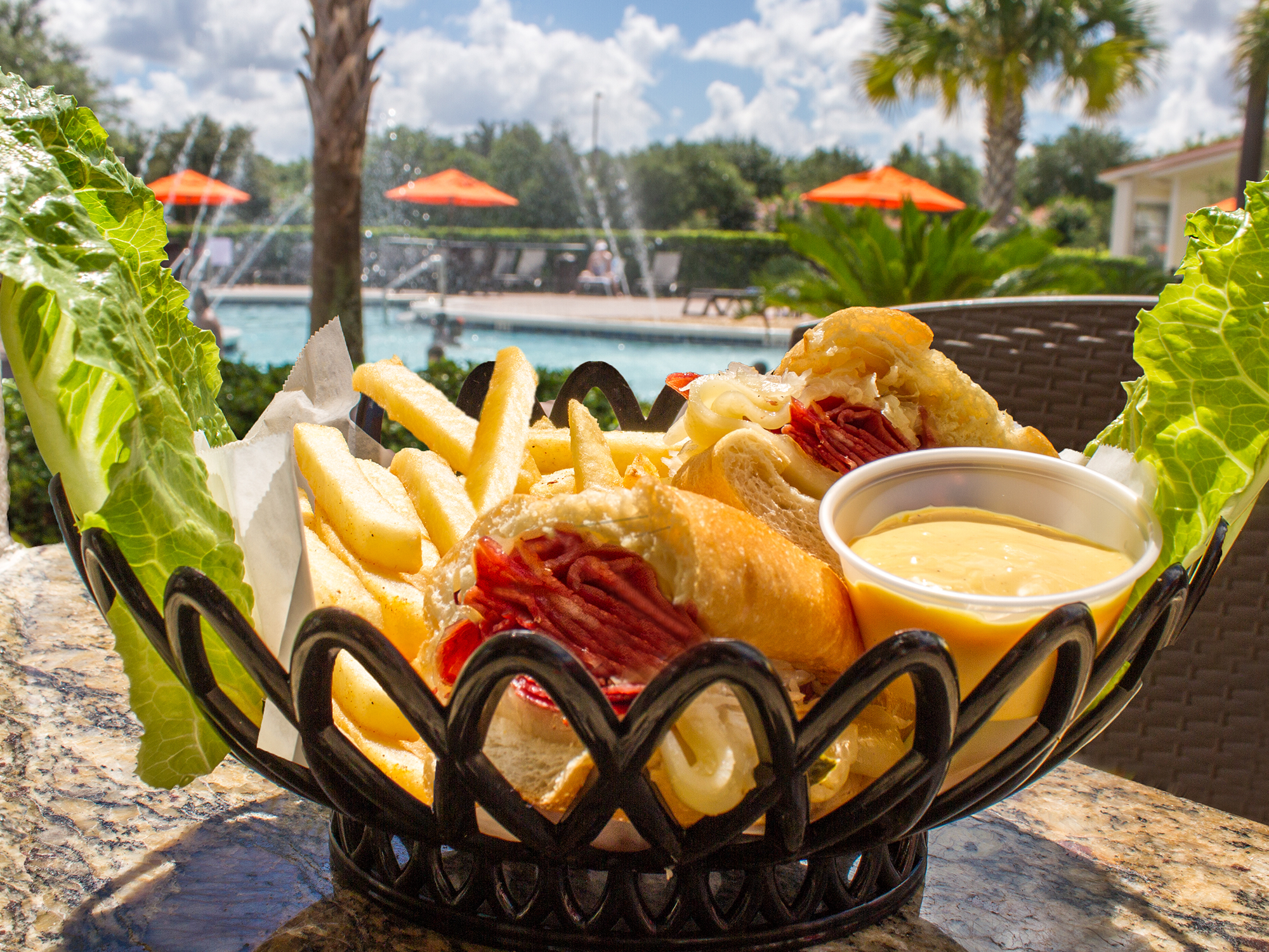 Bowl of food with fries and a sandwich in an outdoor setting with a pool and umbrellas.